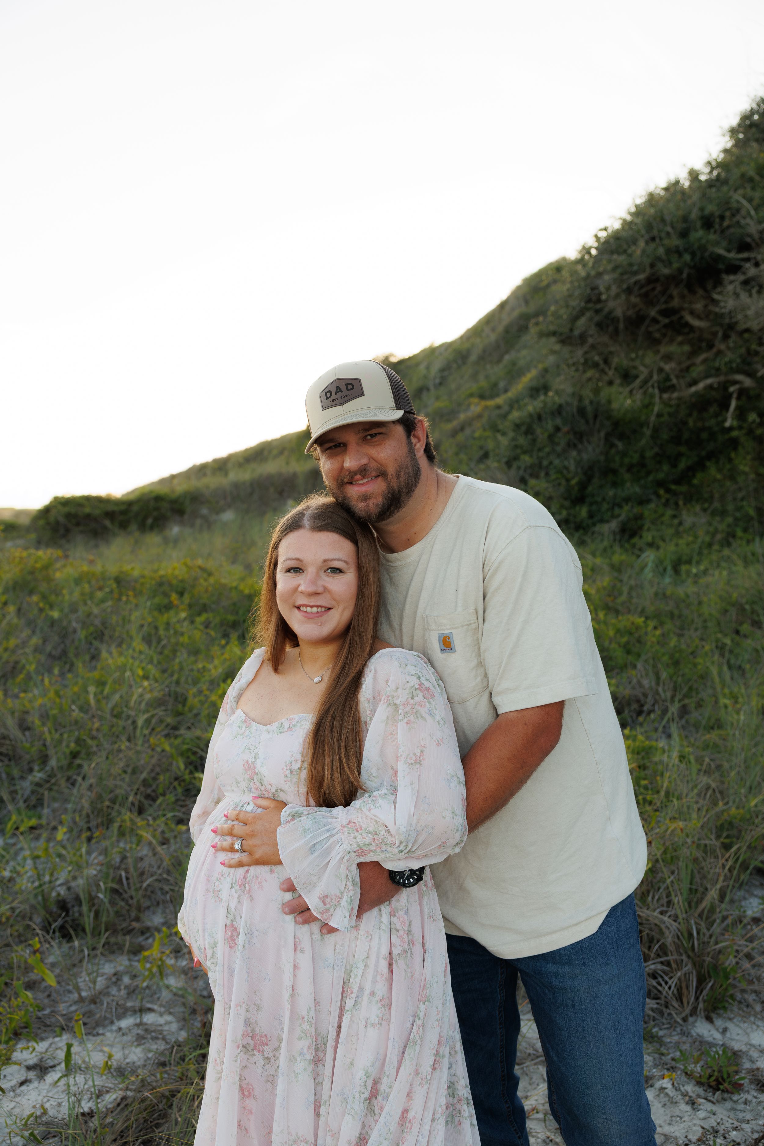 Couple poses outdoors near sandy dunes, woman in floral dress with man in cap embracing her from behind.