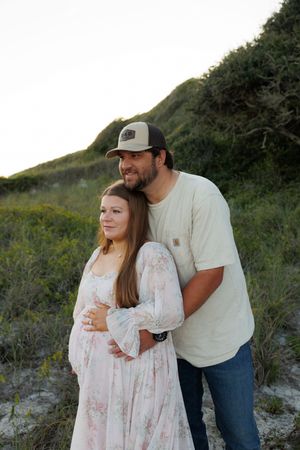 A couple poses outdoors at sunset, woman in floral dress, man in cap embracing her from behind near green dunes.