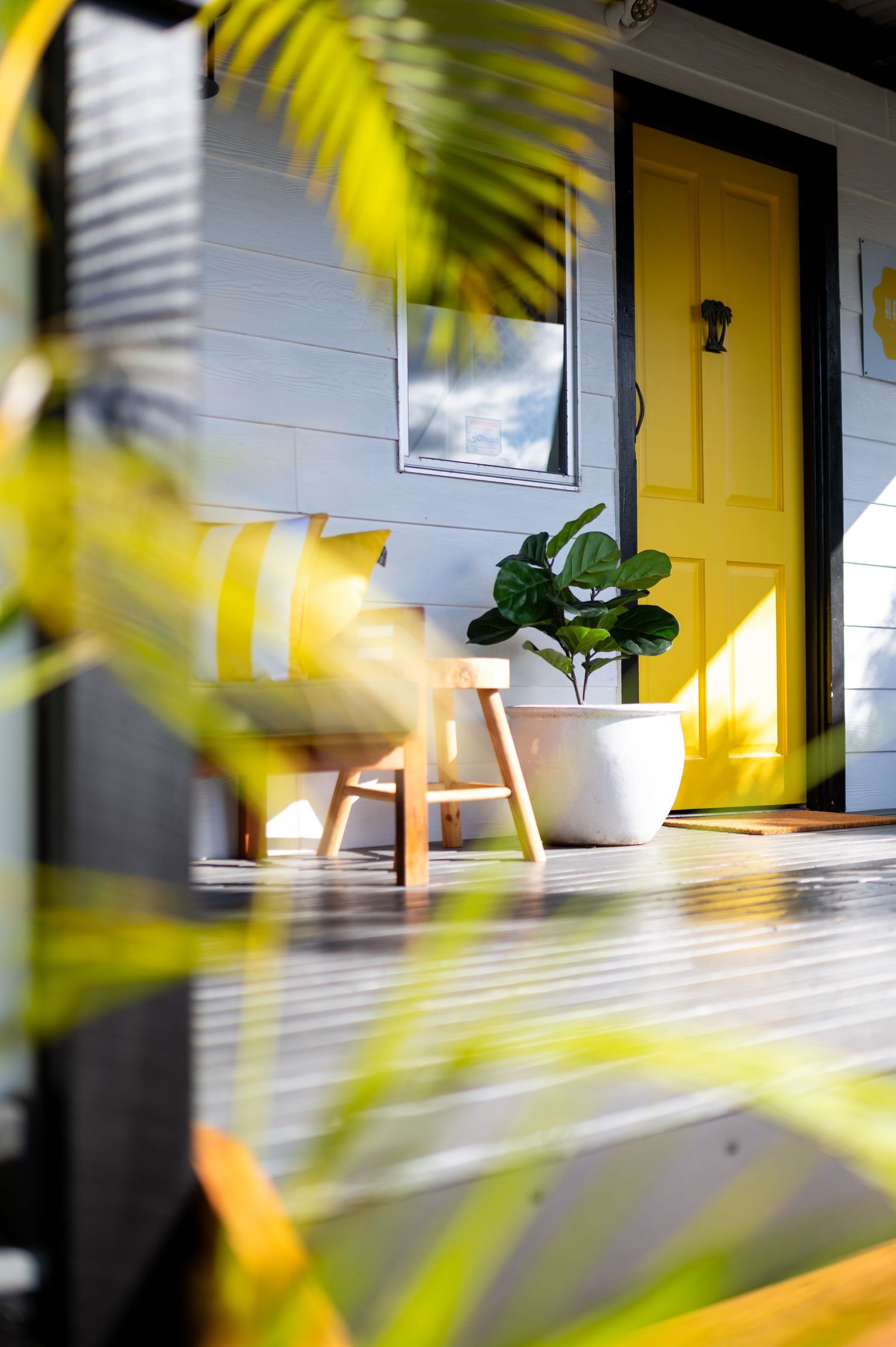 Sunny porch with yellow front door, white potted fiddle leaf fig, wooden stool, and striped cushion on a timber deck.