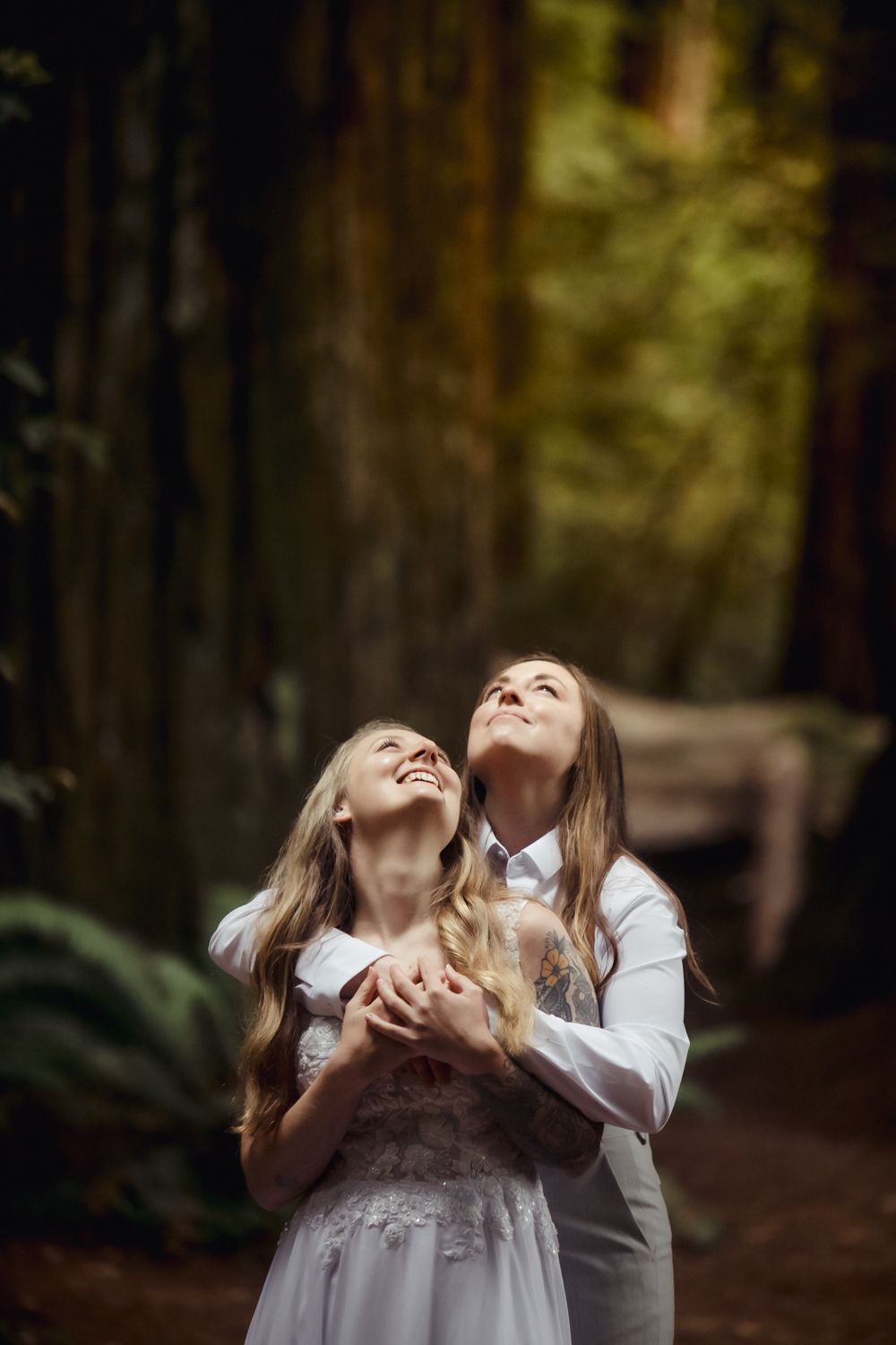 Gay couple standing among towering redwood trees in a lush forest, looking up at the trees