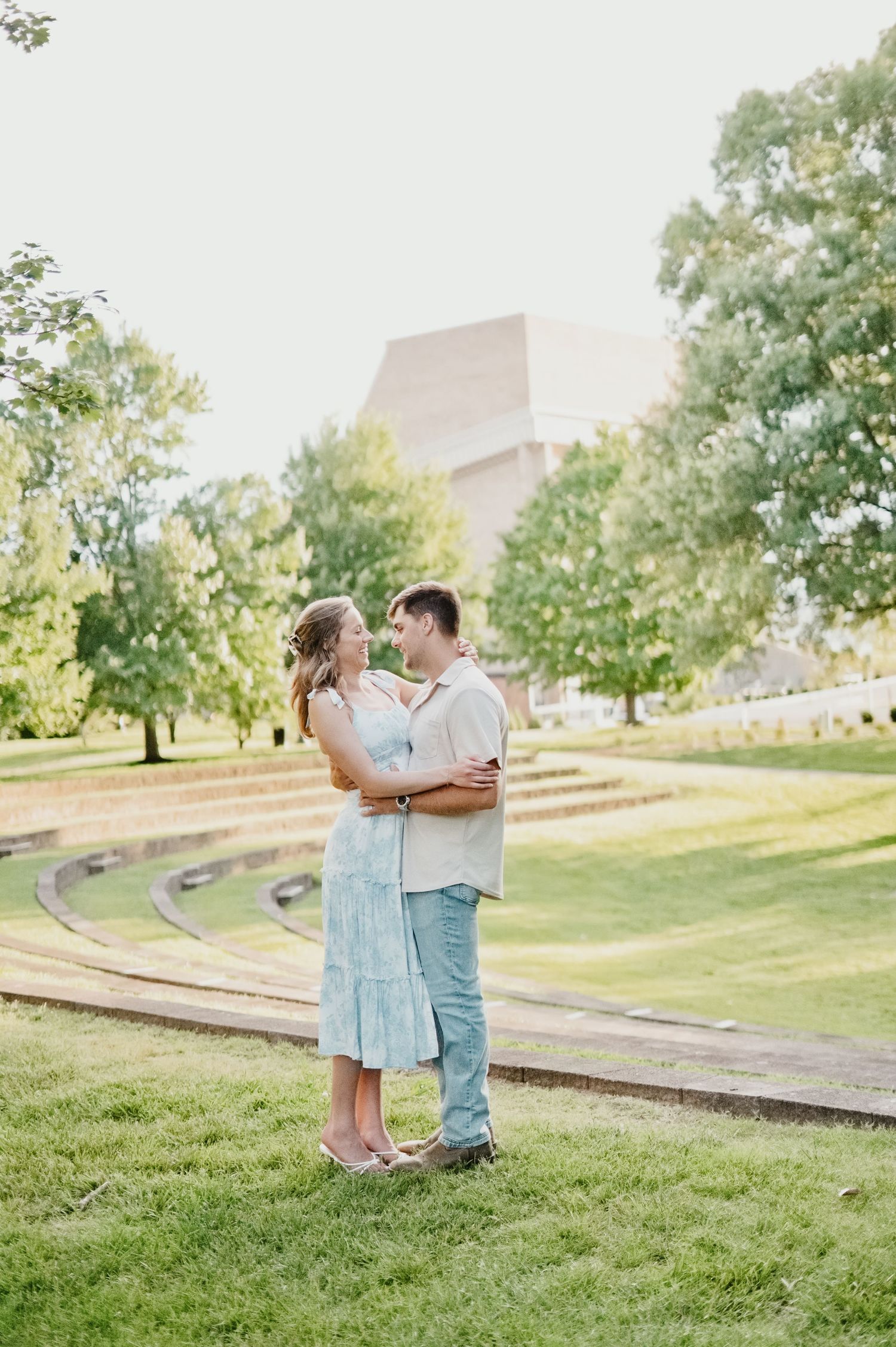 Couple embracing and smiling at each other in a lush green park with trees and a modern building backdrop.