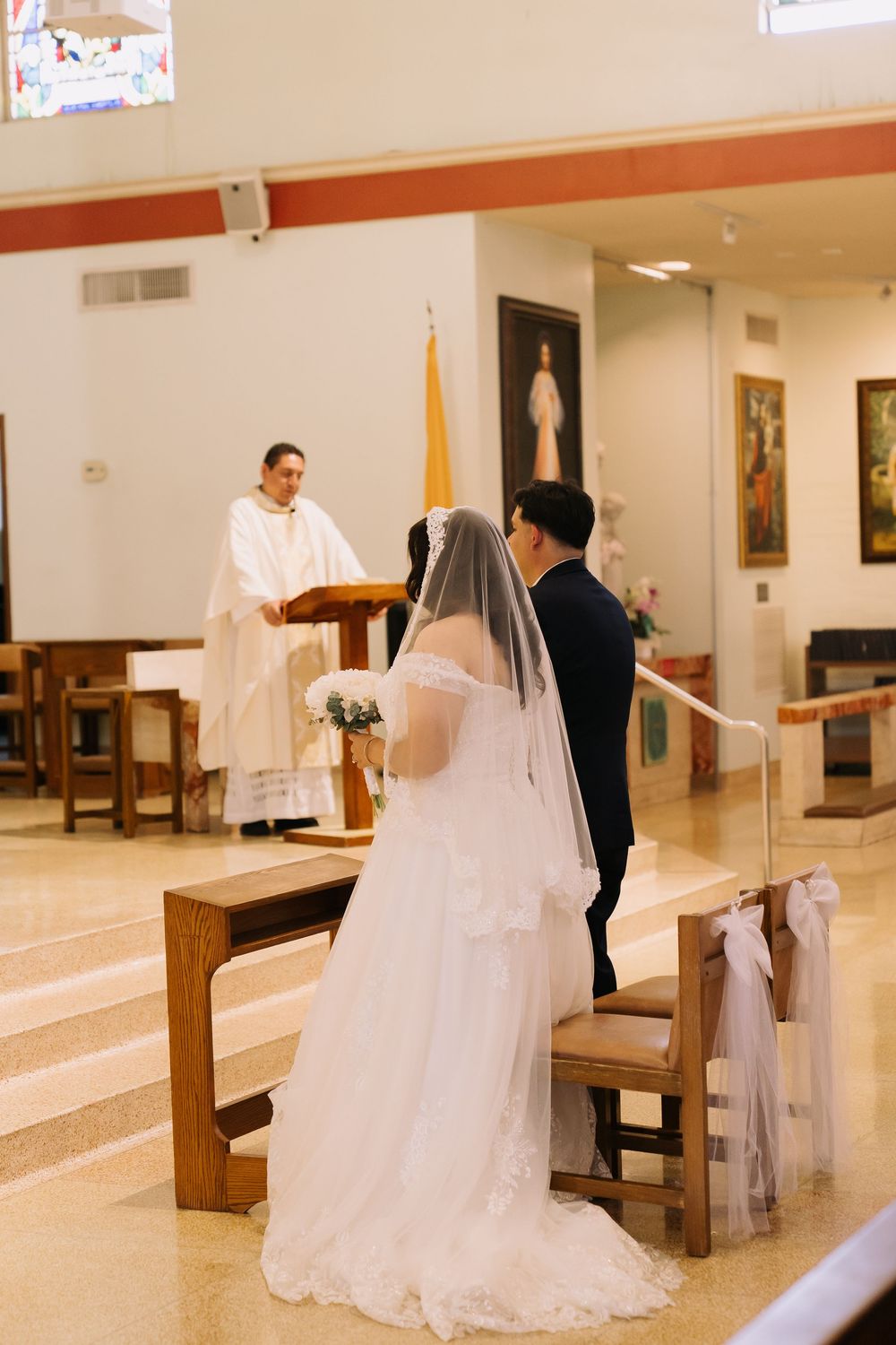 Bride and groom stand at church altar before priest during Catholic wedding ceremony, bride in white gown with veil.