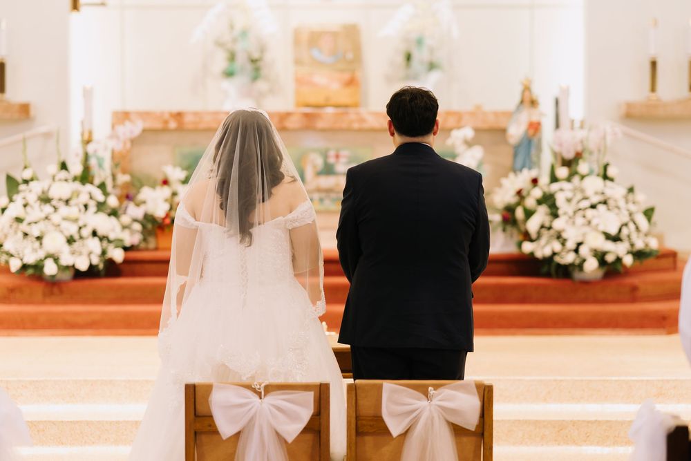 Bride and groom kneeling together at church altar during wedding ceremony with white floral decor.