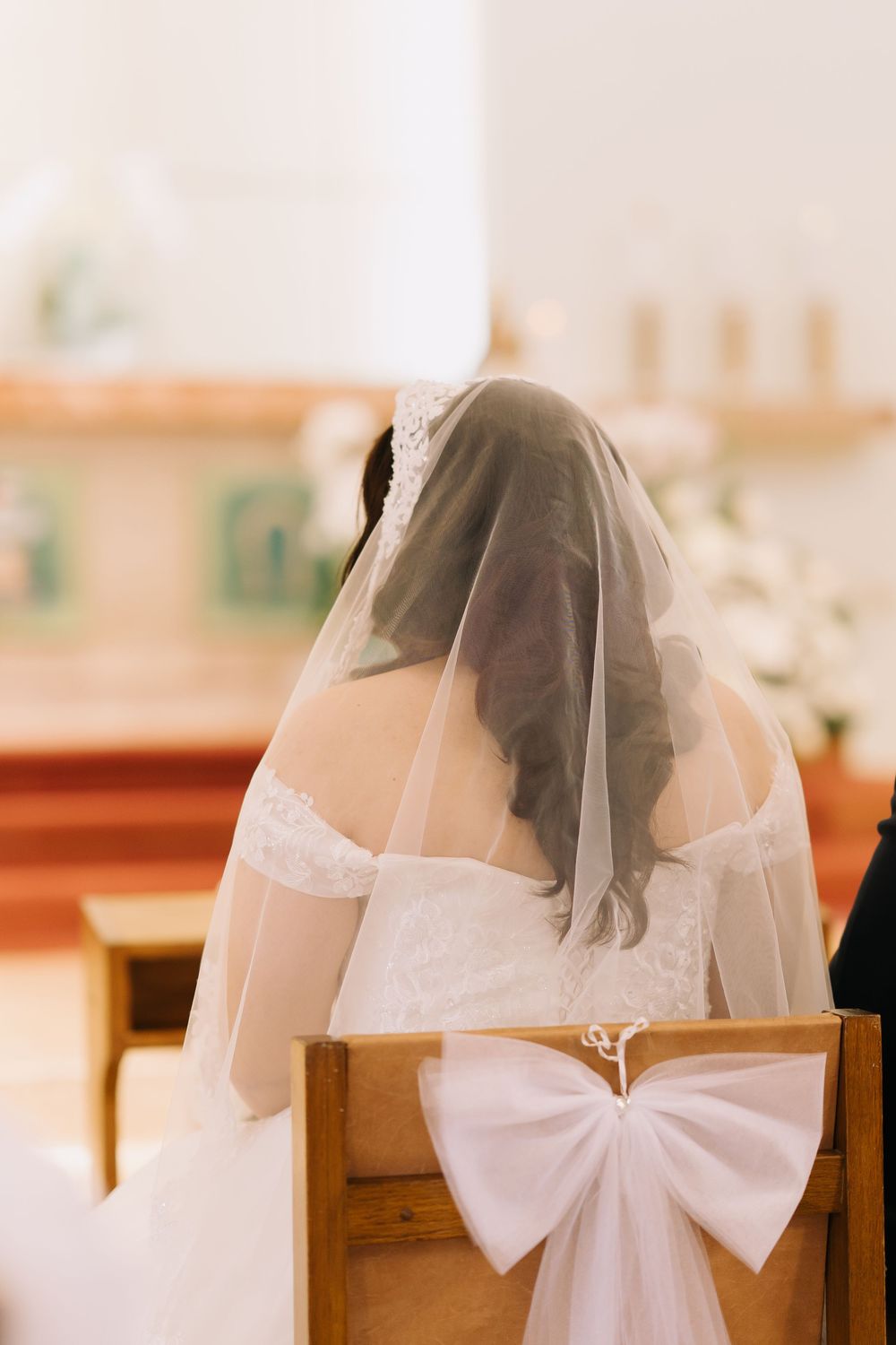 Bride in white lace gown and veil seated in church pew with white bow decoration during wedding ceremony.