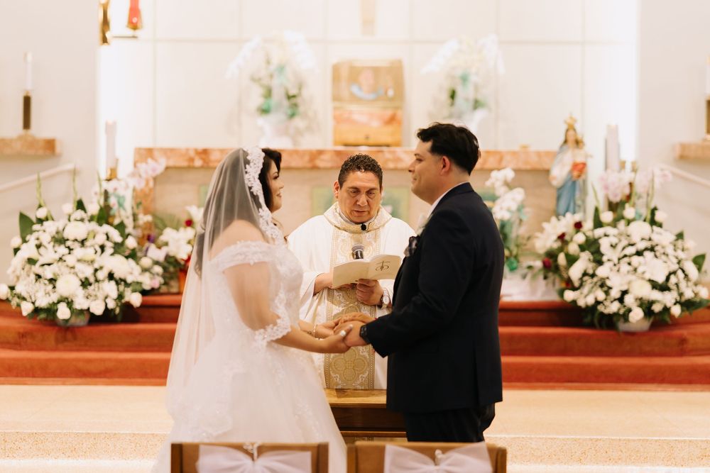 Bride and groom exchange vows at church altar with white floral arrangements and officiant during wedding ceremony.