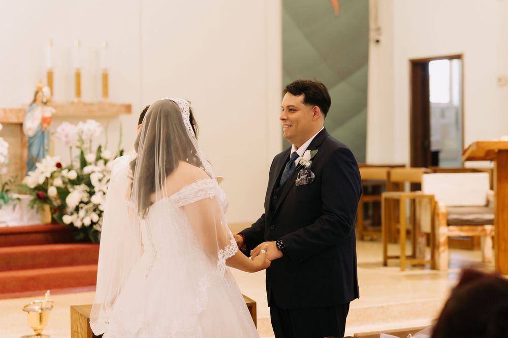 Smiling groom holds bride's hands at church altar during romantic wedding ceremony with floral decorations.
