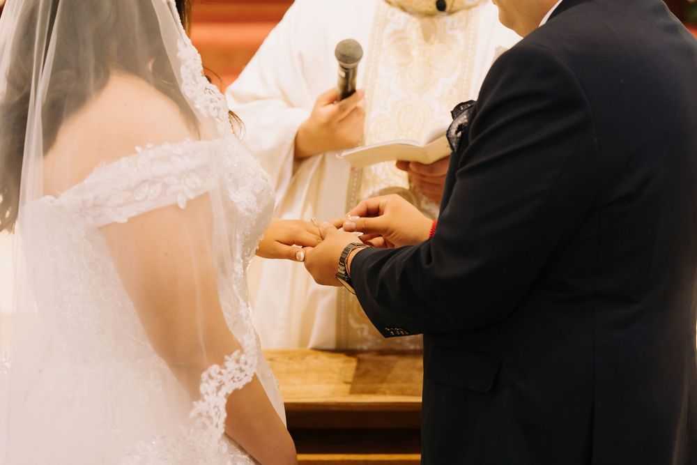 Groom placing wedding ring on bride's finger during church ceremony with officiant holding microphone.