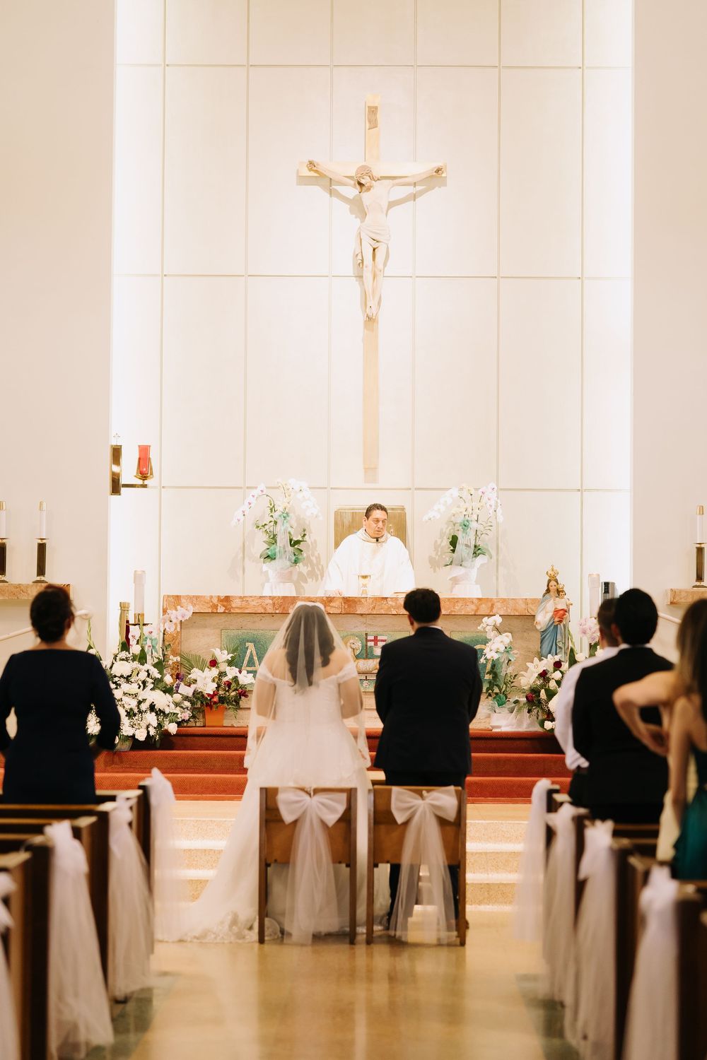 Bride and groom kneeling at church altar during Catholic wedding ceremony with priest and crucifix.