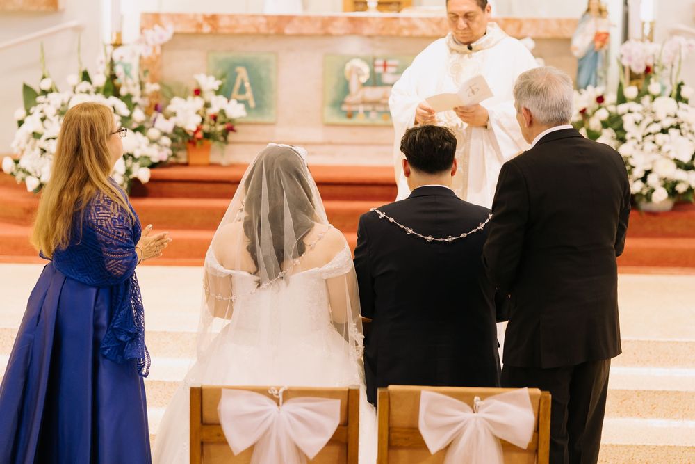 A bride and groom kneel before a priest at a church altar during a traditional wedding ceremony with floral decorations.