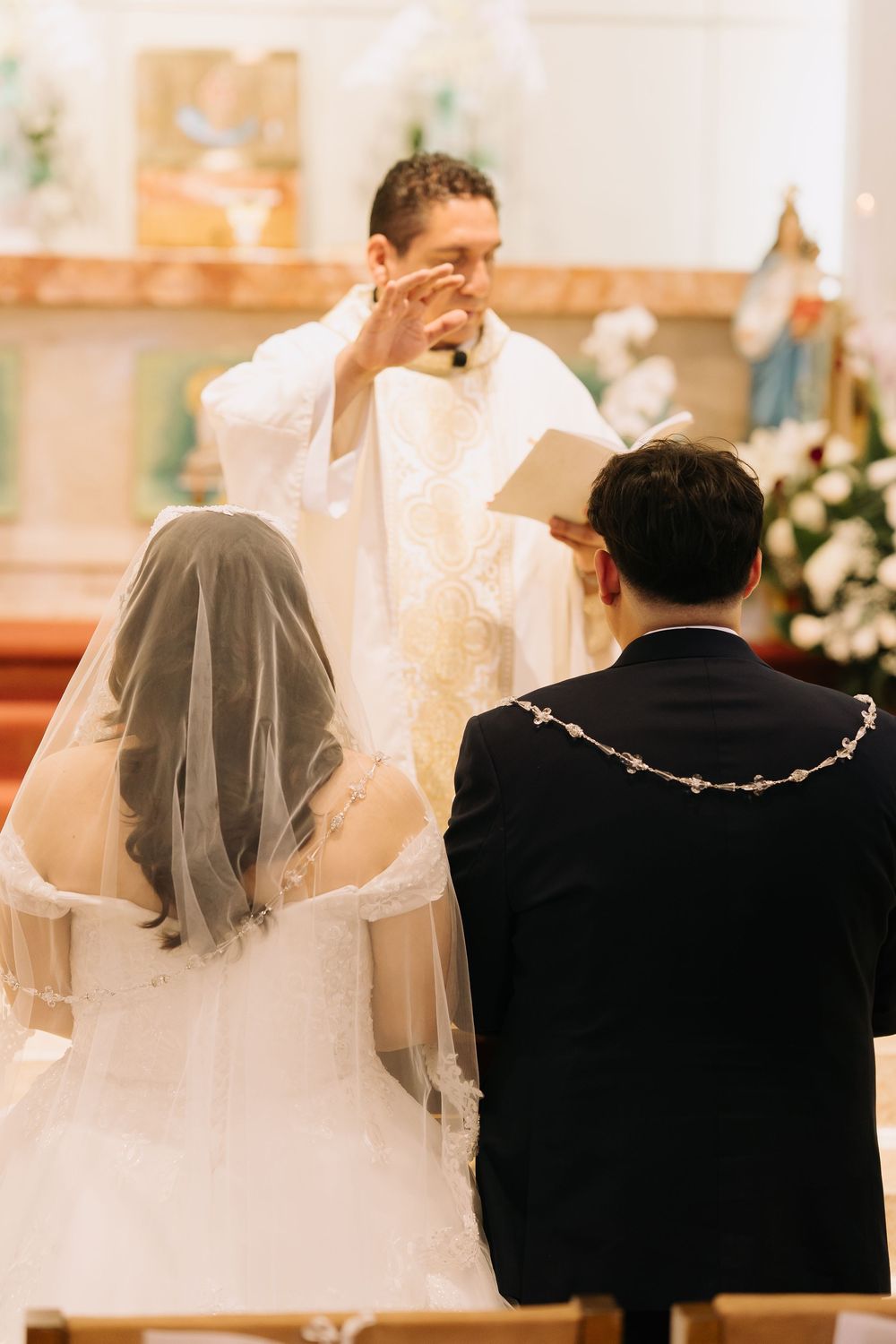 Priest officiating a Catholic wedding ceremony with bride in white veil and groom in black suit at church altar.