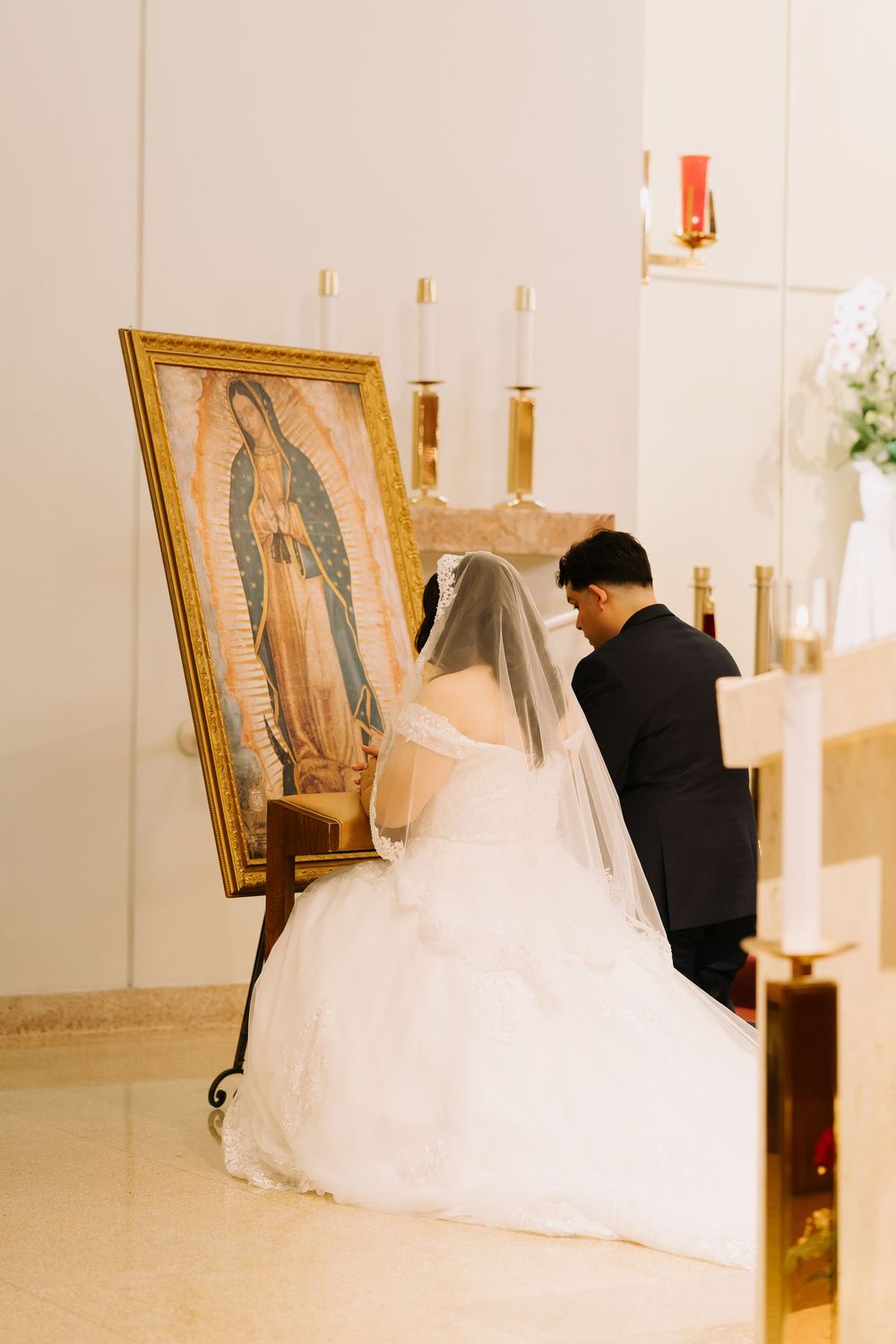 Bride and groom kneeling in prayer before Our Lady of Guadalupe painting during Catholic wedding ceremony.
