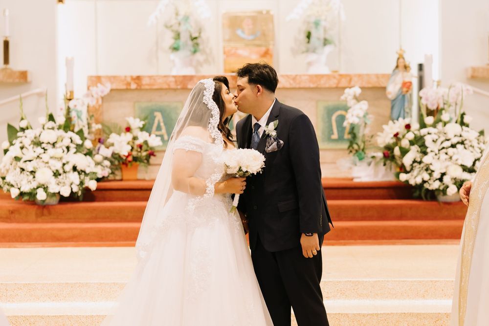 Bride and groom share first kiss at church altar surrounded by white floral arrangements.