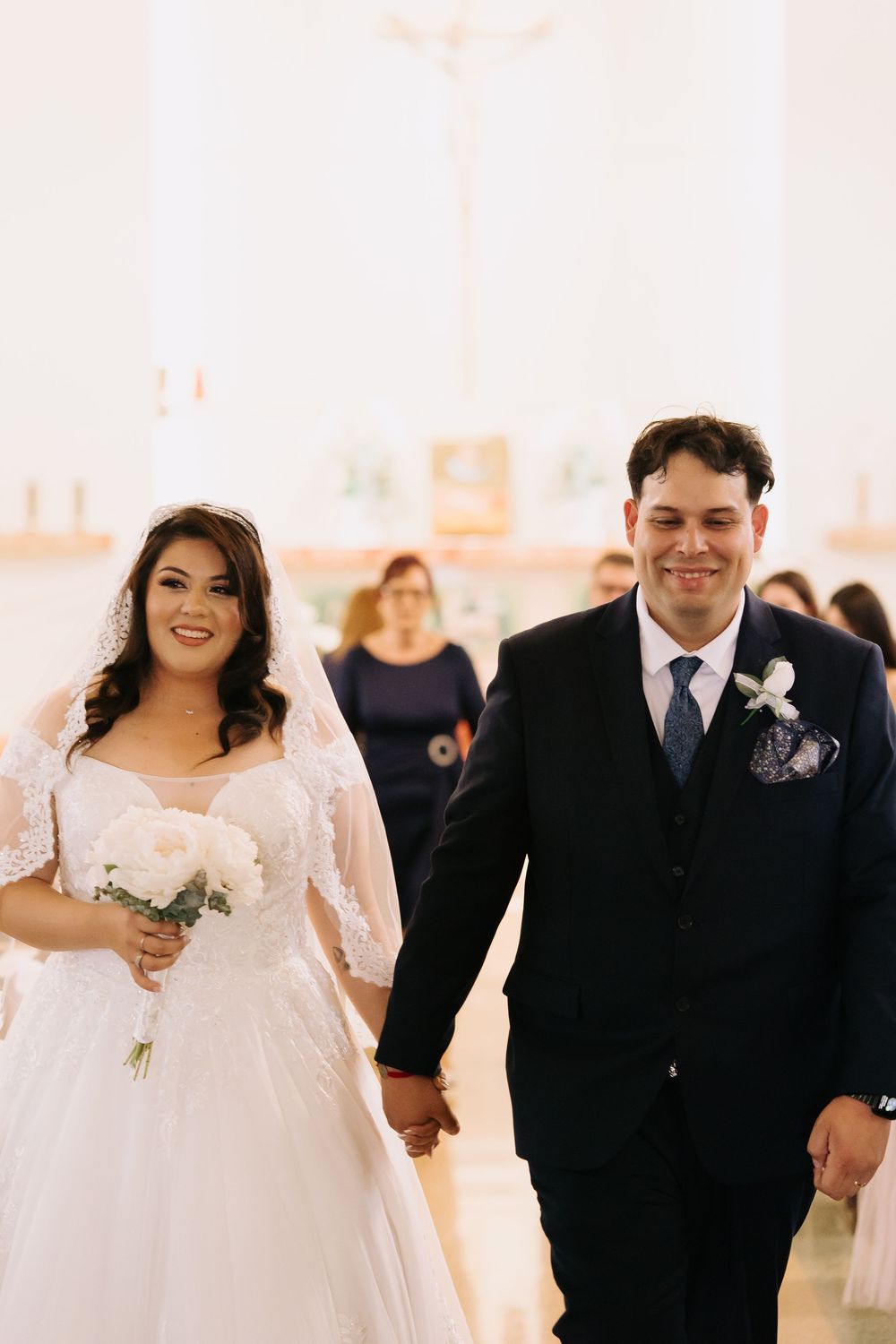 Bride and groom walk hand-in-hand down church aisle, bride in lace gown holding white bouquet, groom in navy suit.