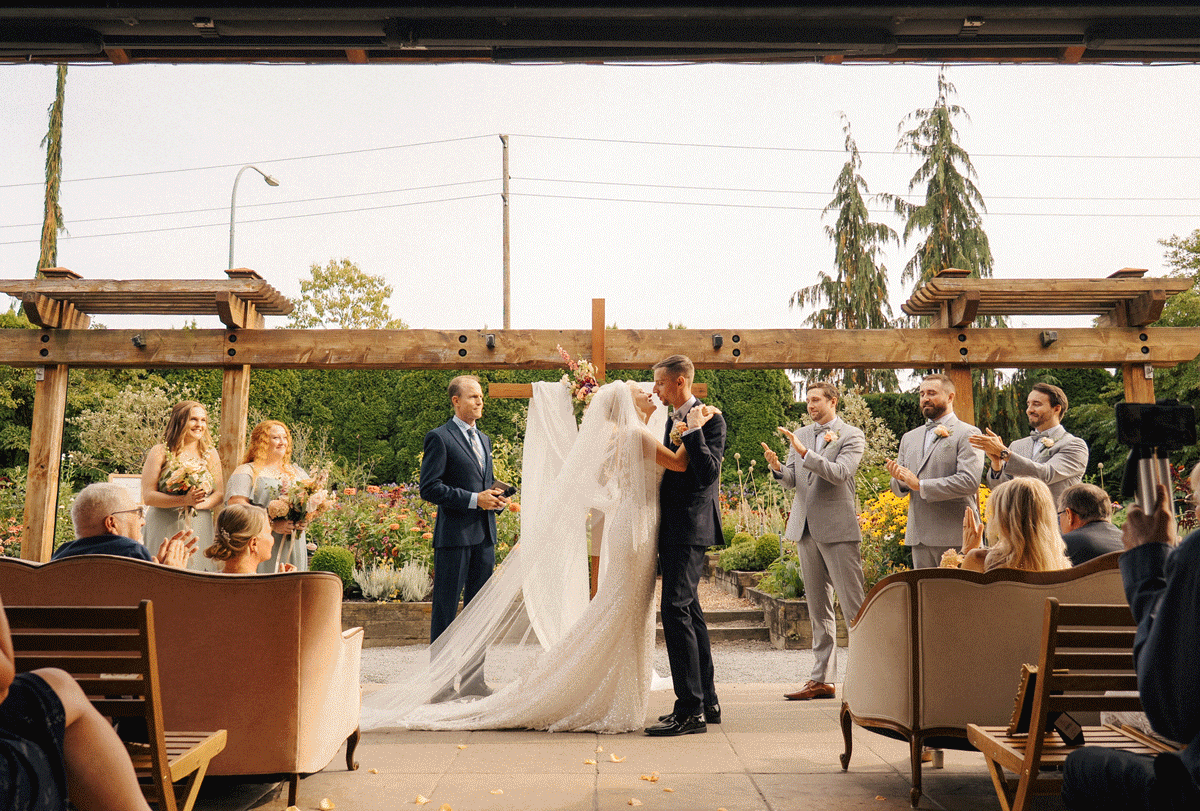 Outdoor wedding ceremony under a pergola with bride, groom, and wedding party surrounded by lush greenery.