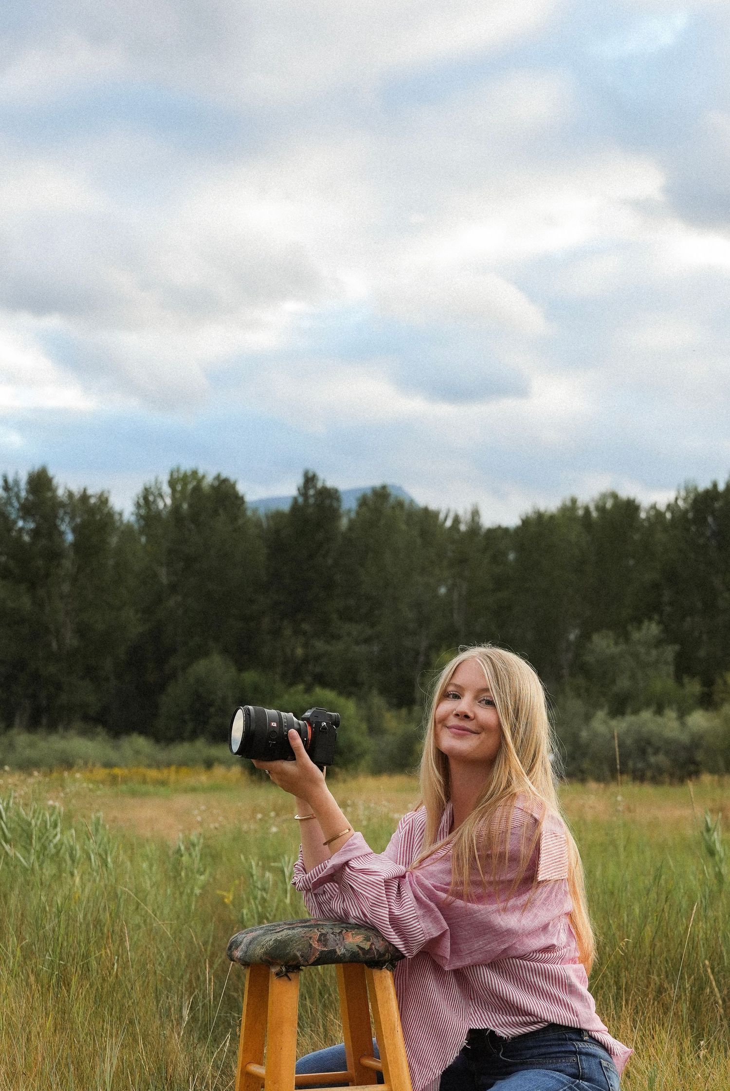 Blonde woman in pink off-shoulder top holds camera while sitting on stool in scenic green meadow with trees and cloudy sky.