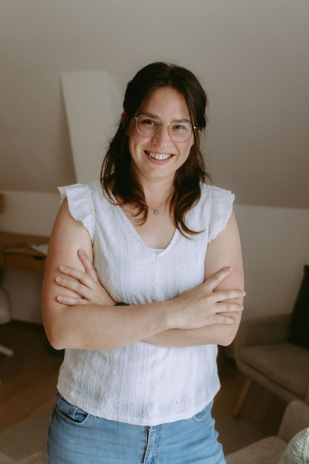 Smiling woman with glasses and crossed arms wearing a white sleeveless top and jeans in a cozy indoor setting.