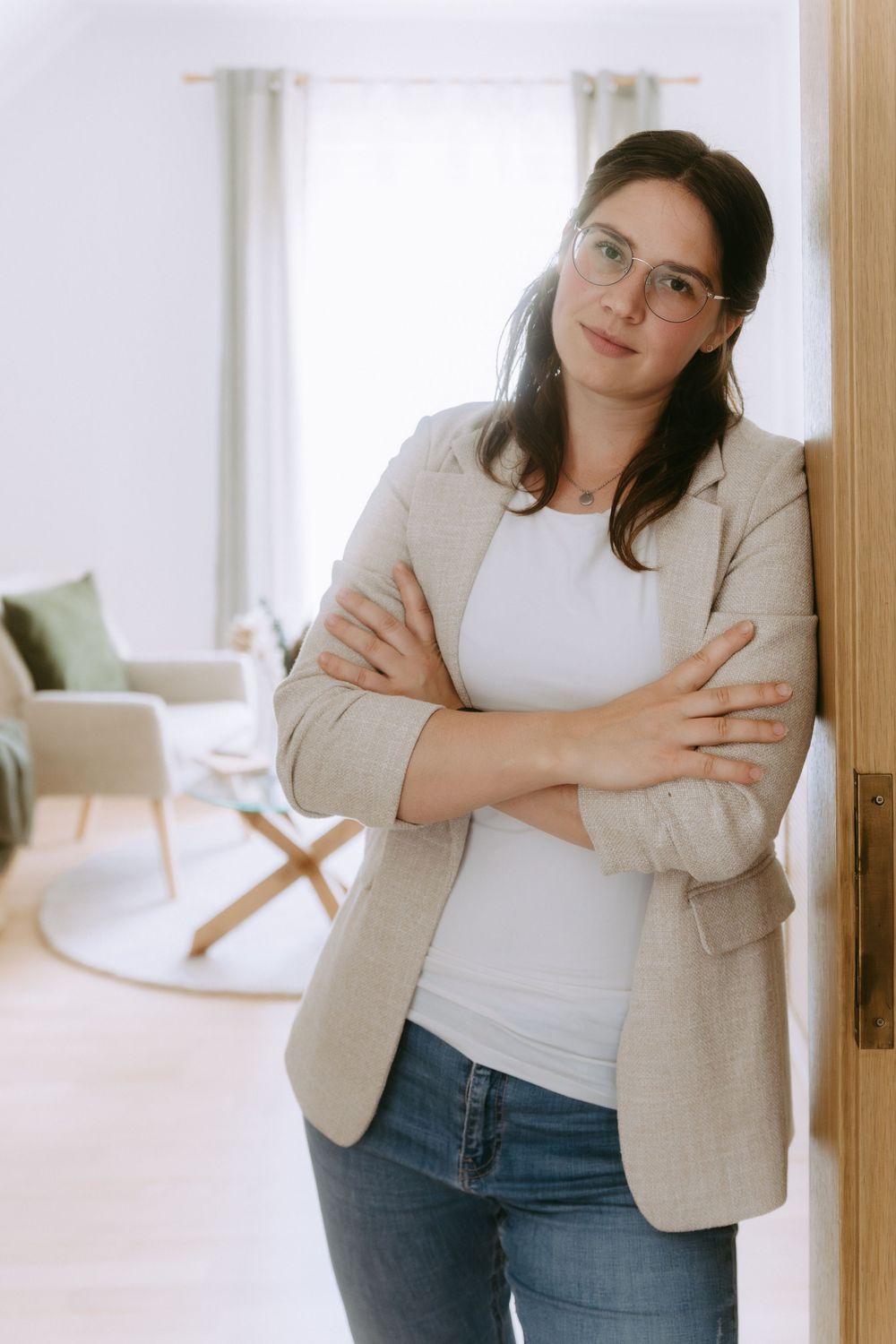 Woman in glasses and blazer leaning against doorframe with arms crossed in modern living room.