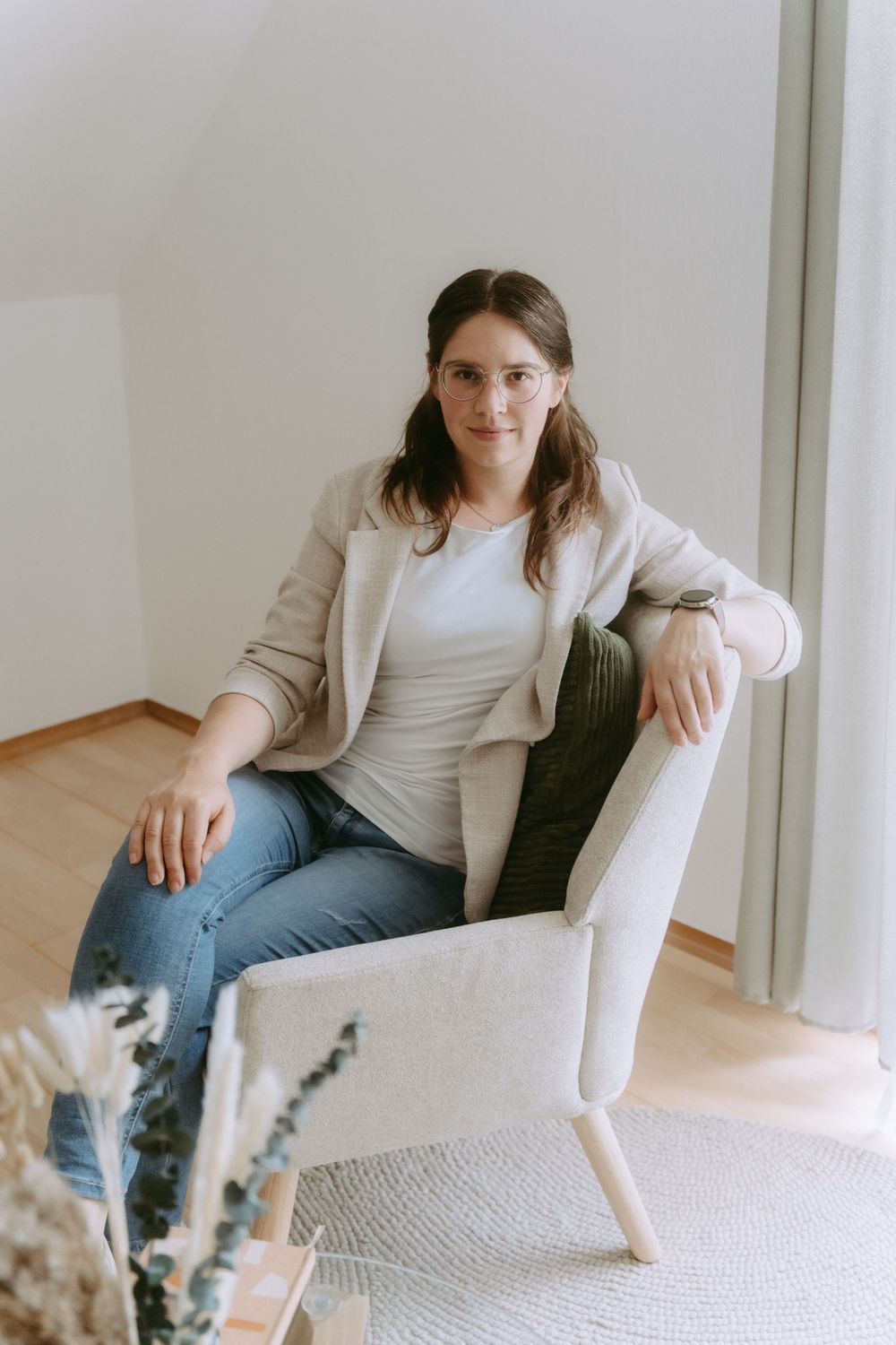 Woman in beige blazer and jeans sitting casually in a light modern armchair near a curtained window.