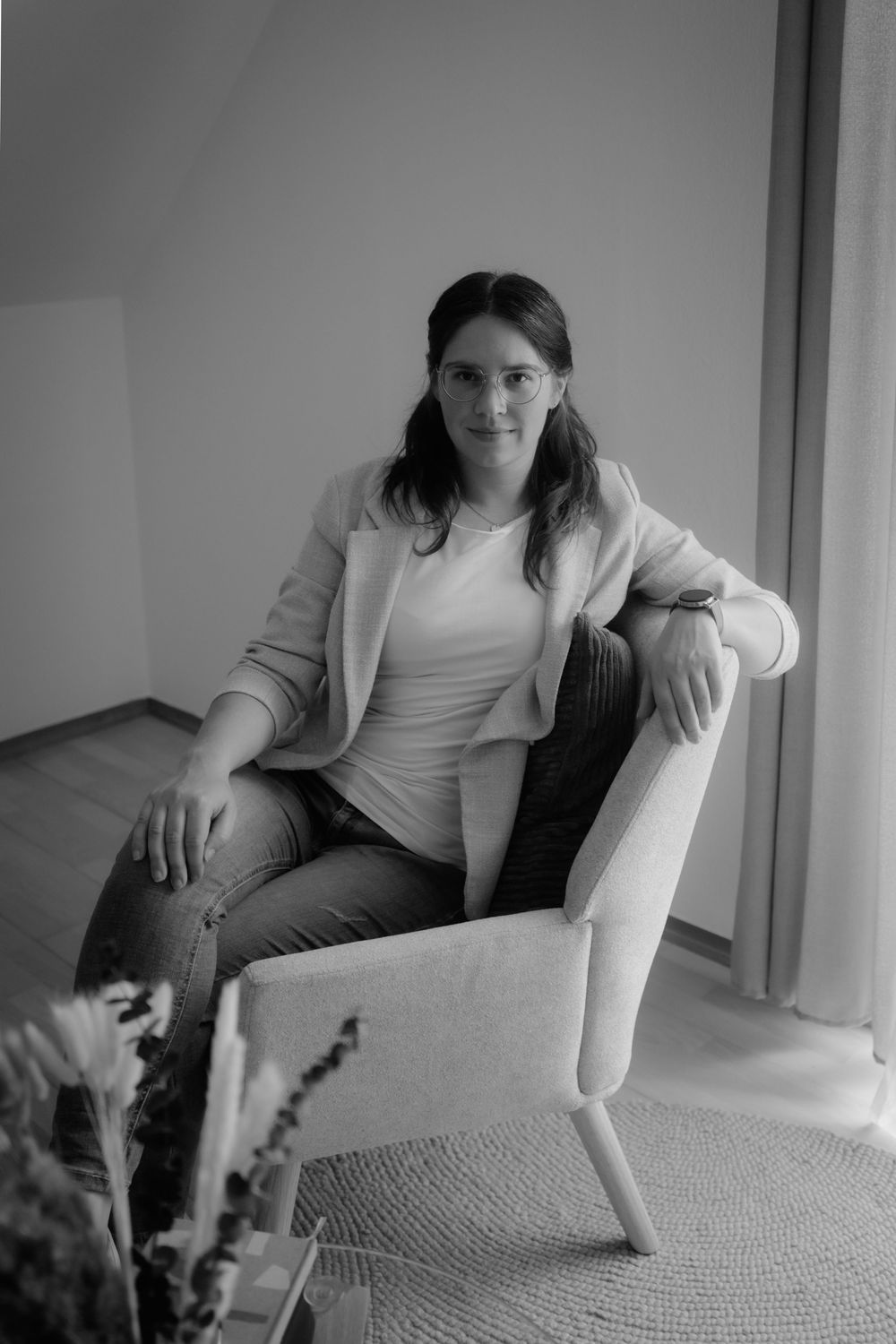 Black and white portrait of a woman in blazer sitting relaxed in a modern armchair near a window.