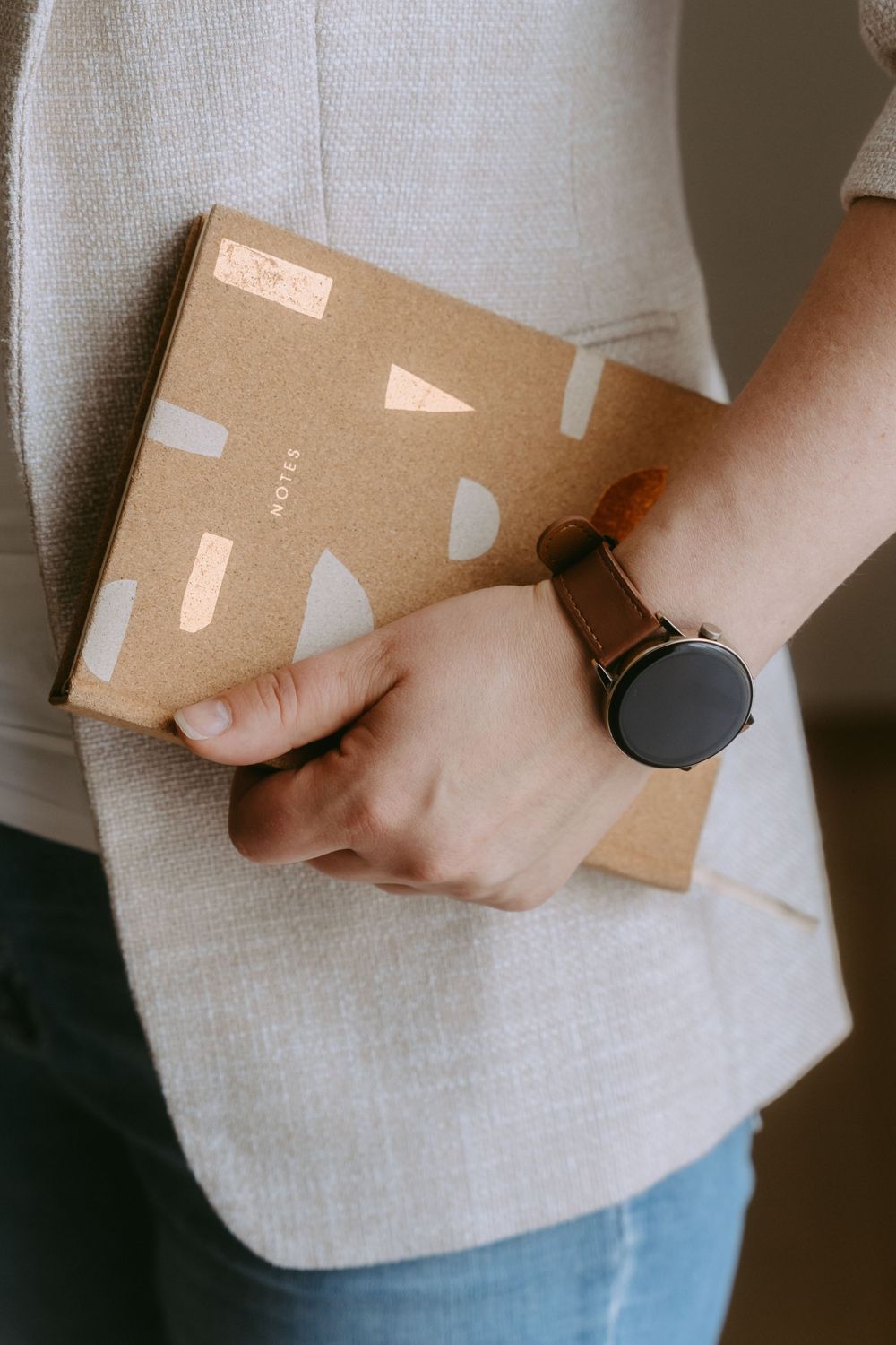 Person holding a kraft paper notebook with geometric shapes, wearing a black minimalist watch and light gray blazer.