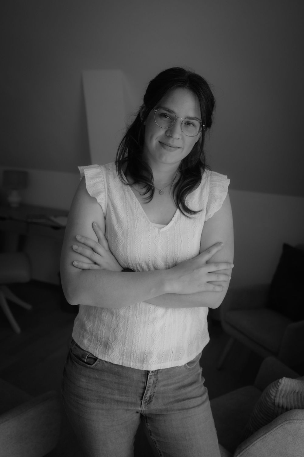 Black and white portrait of a smiling woman with glasses, arms crossed, wearing a white top and jeans indoors.