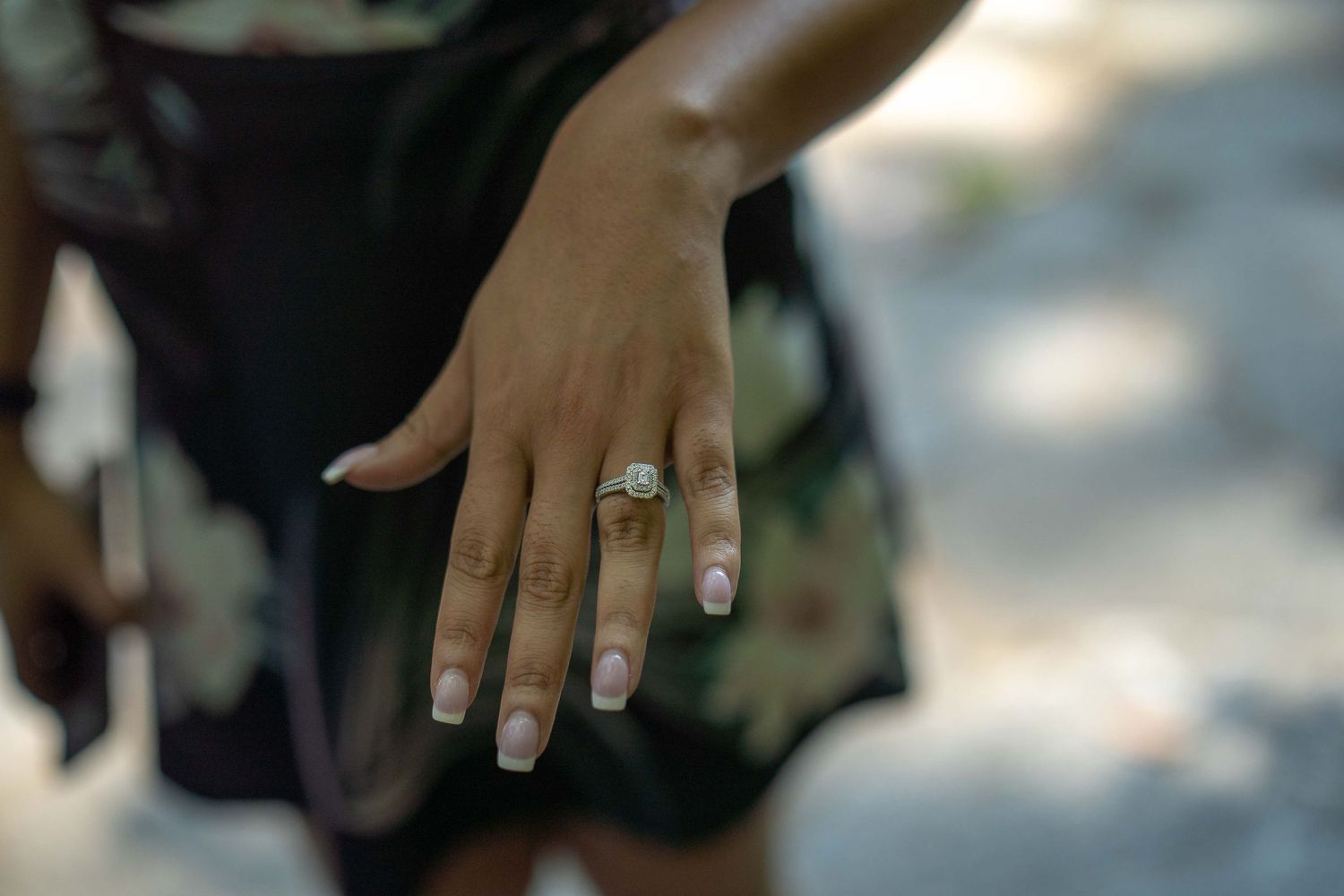 Close-up of a woman's hand displaying a diamond engagement ring with elegant nail extensions outdoors.