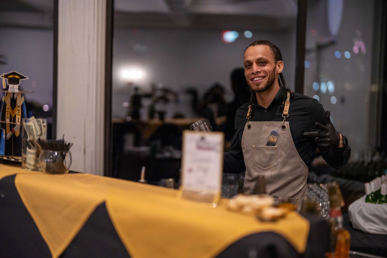 Smiling barista in apron stands behind counter at coffee bar with yellow tablecloth in modern cafe.