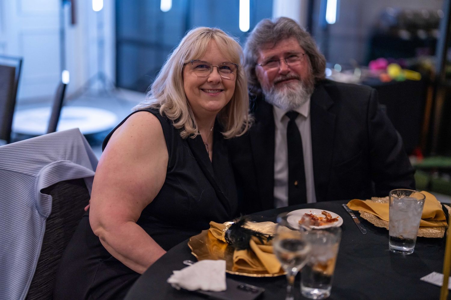 Couple smiling together at a formal dinner event, dressed in black attire, seated at a table with gold napkins.
