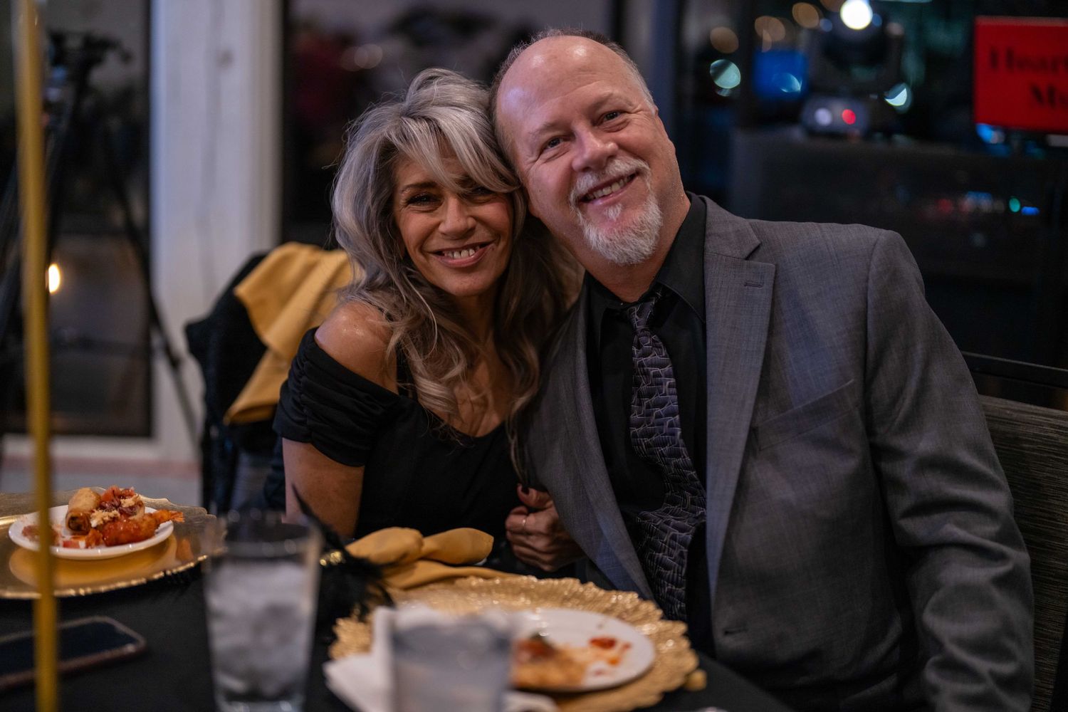 Smiling couple posing together at a restaurant dinner table, man in gray blazer and woman in black off-shoulder top.