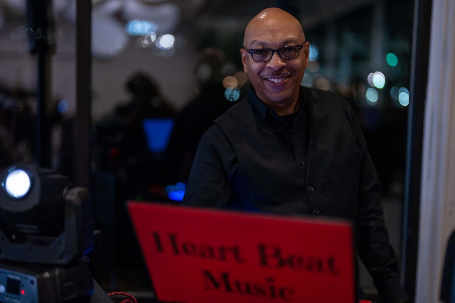 Smiling DJ stands behind Heart Beat Music sign at an evening event with stage lighting equipment.