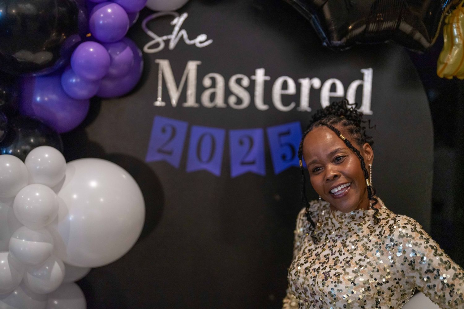 Woman smiling at She Mastered 2025 graduation celebration with purple, white, and black balloons.