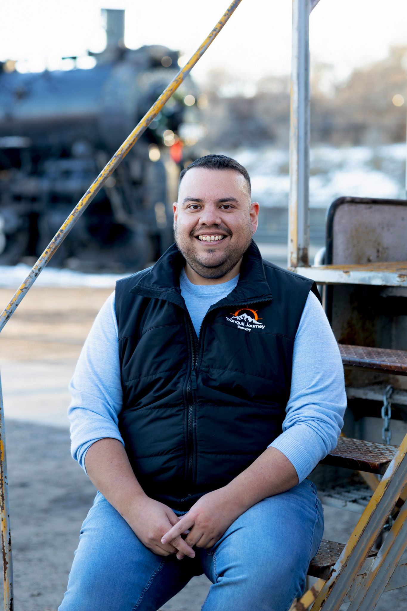 Smiling man in black vest sitting outdoors near vintage train equipment on a sunny day.