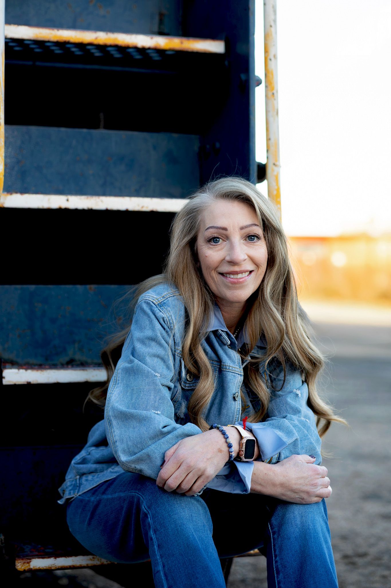 Blonde woman in denim jacket sitting outdoors near blue metal steps, smiling warmly.