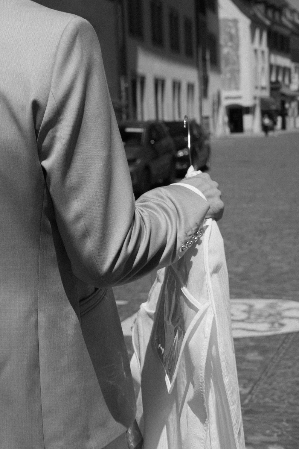 Black and white photo of a groom in a suit holding a bride's white wedding dress on a cobblestone street.