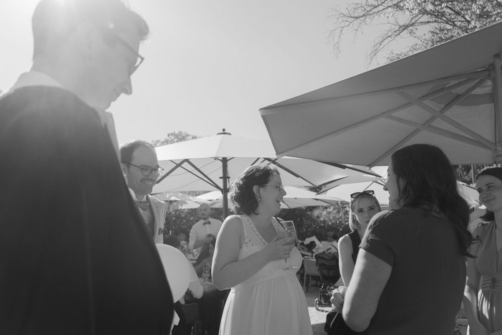 Black and white wedding reception photo with guests mingling outdoors under umbrellas in bright sunlight.