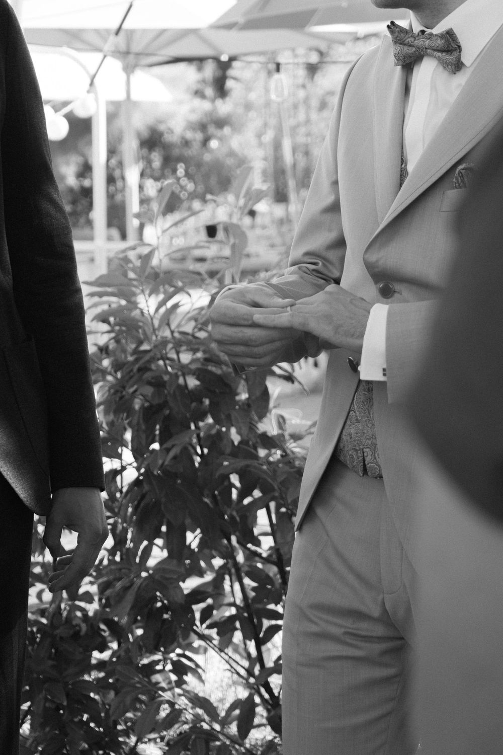Black and white photo of two men in suits holding hands at a wedding ceremony outdoors.