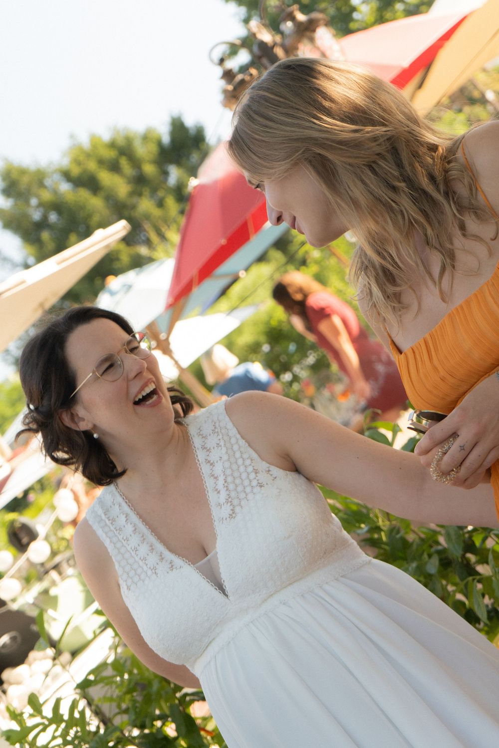 Two women laughing together outdoors at a wedding celebration, bride in white dress with colorful umbrellas in background.