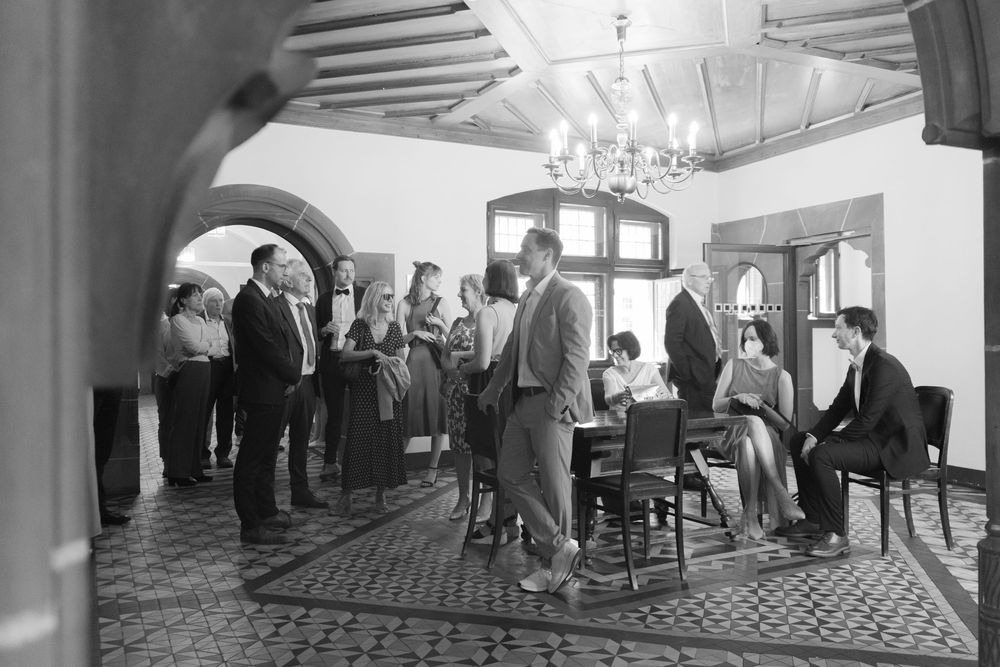 Black and white photo of elegantly dressed people socializing in a chandelier-lit room with arched doorways.