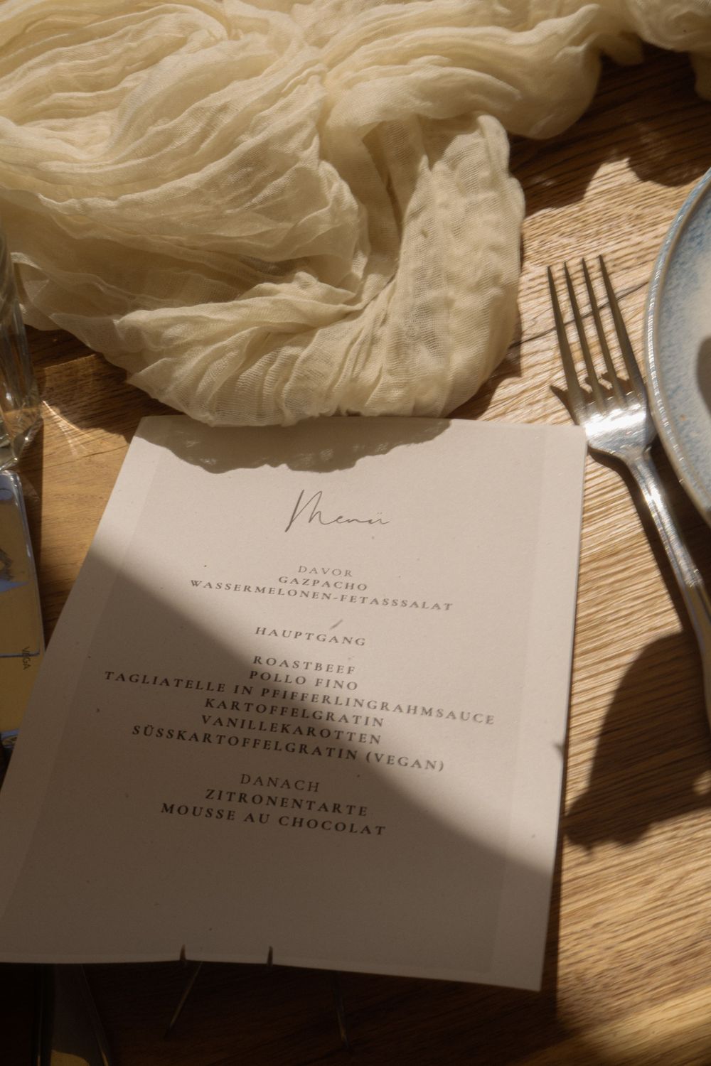 Elegant wedding menu card on wooden table with fork, plate, and delicate cream fabric at a reception.