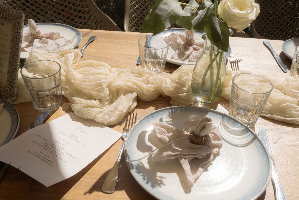 Elegant table setting with white plates, folded napkins, glassware, floral centerpiece, and a decorative cheesecloth table runner.