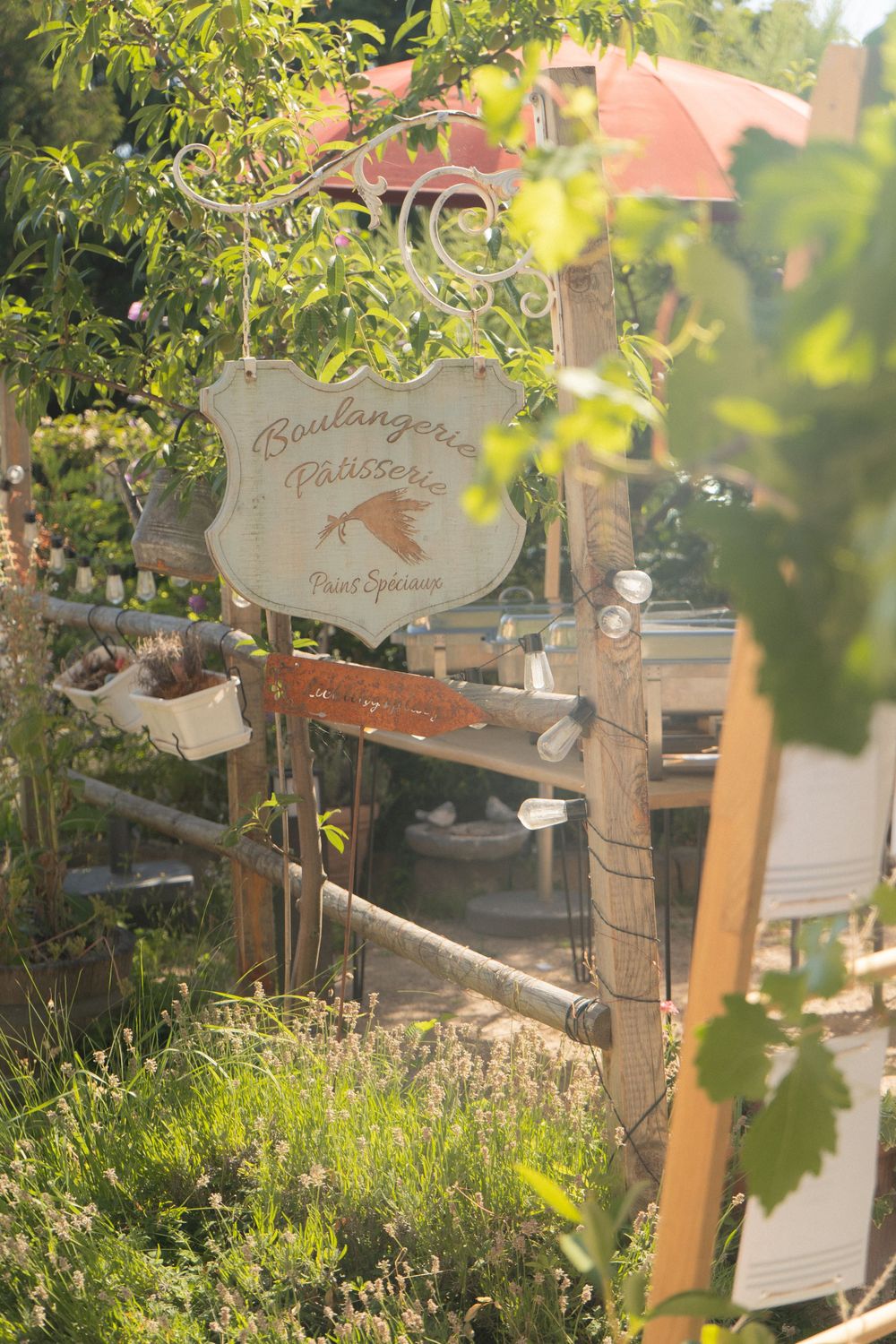 Rustic Boulangerie Patisserie sign hanging in a lush garden surrounded by green plants in warm sunlight.