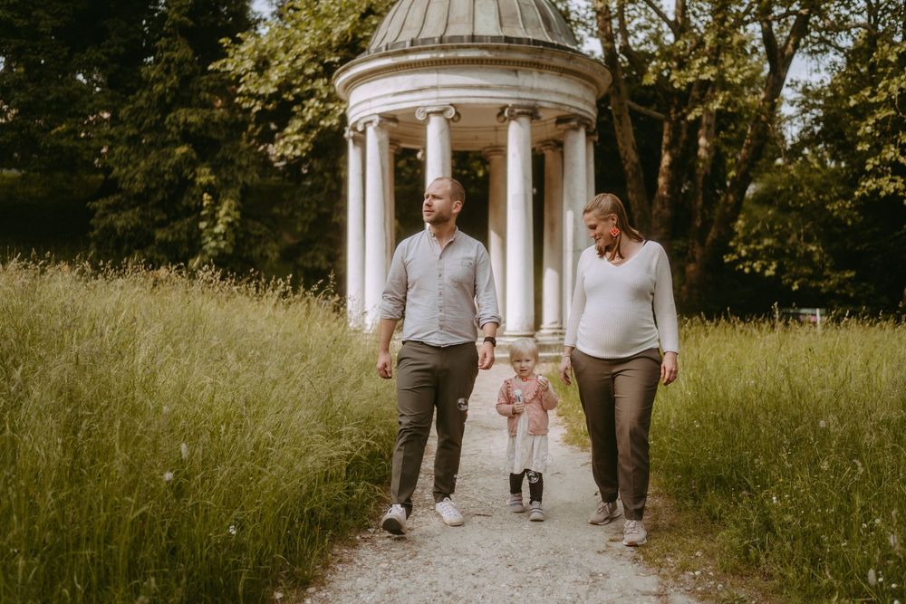 Family of three walking on a garden path, pregnant mother and father holding toddler's hands near a white gazebo.