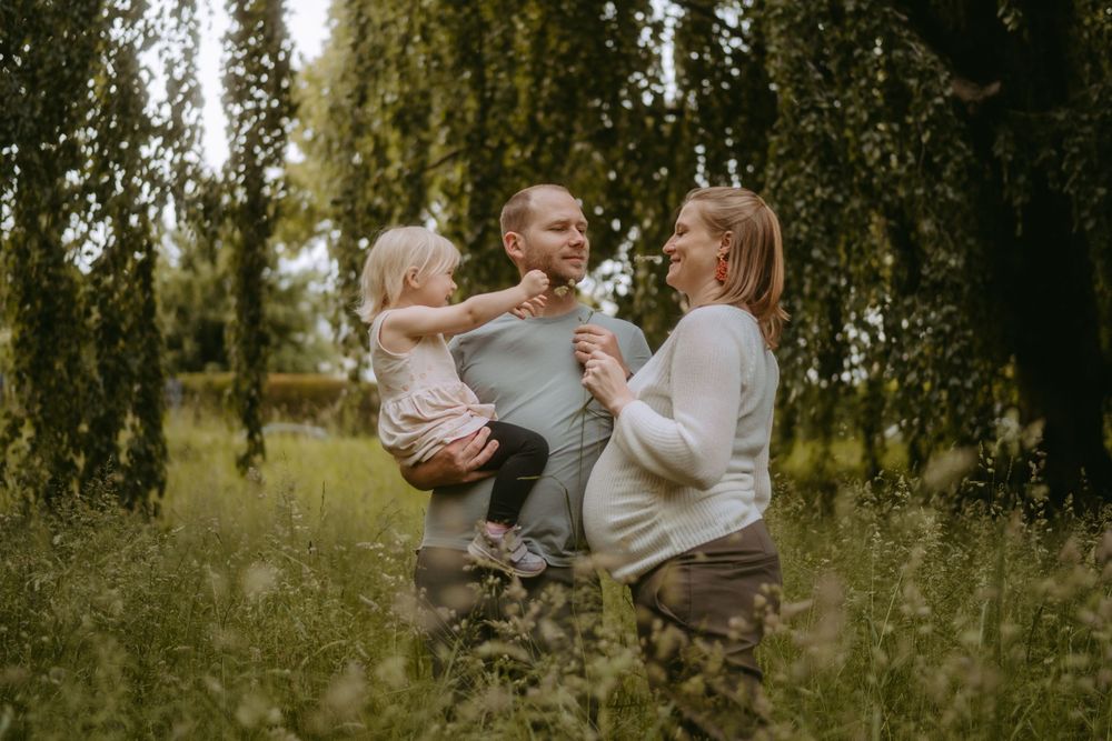 Pregnant woman and man holding toddler girl in lush green park, happy family maternity photo outdoors.