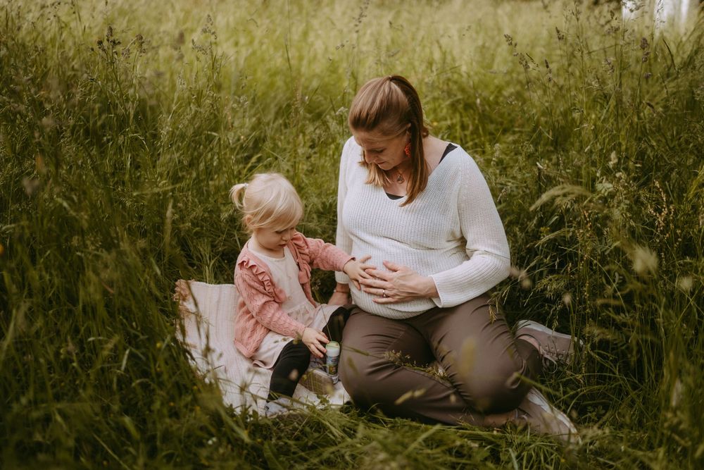 Pregnant woman kneeling in tall grass with young blonde child touching her baby bump during outdoor maternity photo session.