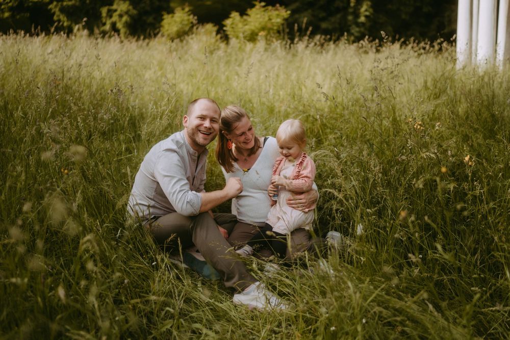 Family of three smiling together in a lush green meadow, parents crouching beside their baby on a sunny day.