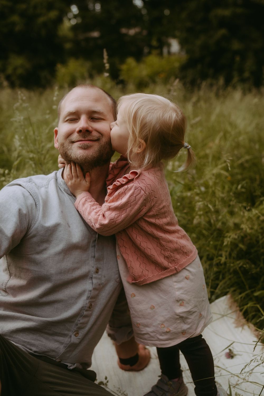 A bald man smiles as a young blonde toddler girl kisses his cheek in a lush green meadow.