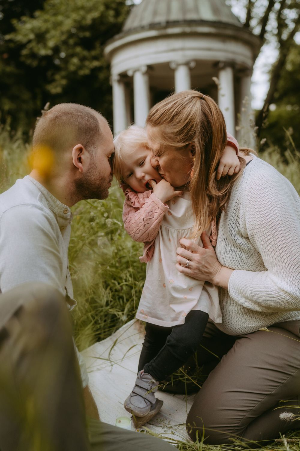 A loving family shares a tender moment outdoors, with parents kissing their young daughter near a classical garden temple.