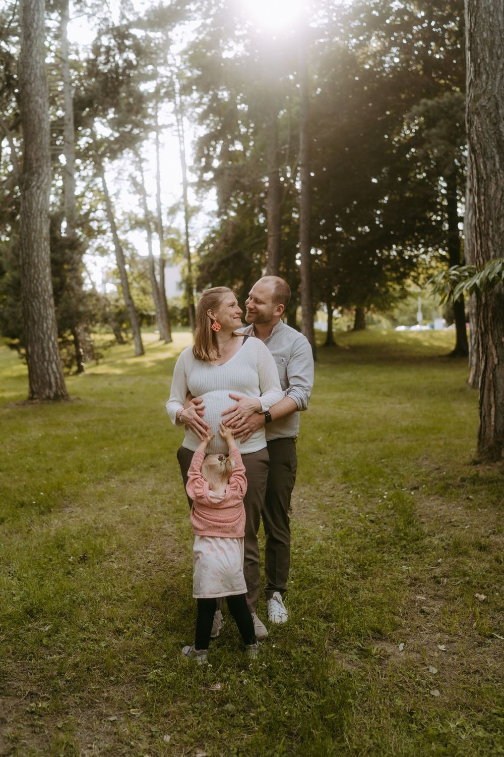Pregnant couple embraces in sunlit park while young child hugs mother's belly outdoors.