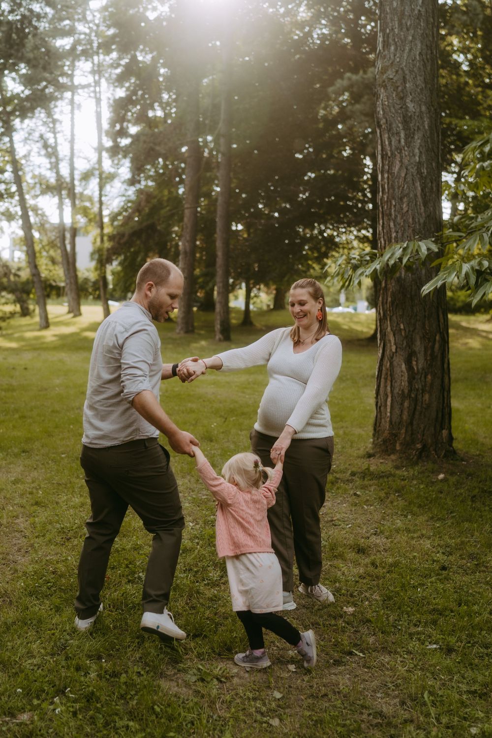 Pregnant woman and man hold hands with toddler girl dancing in sunny park.