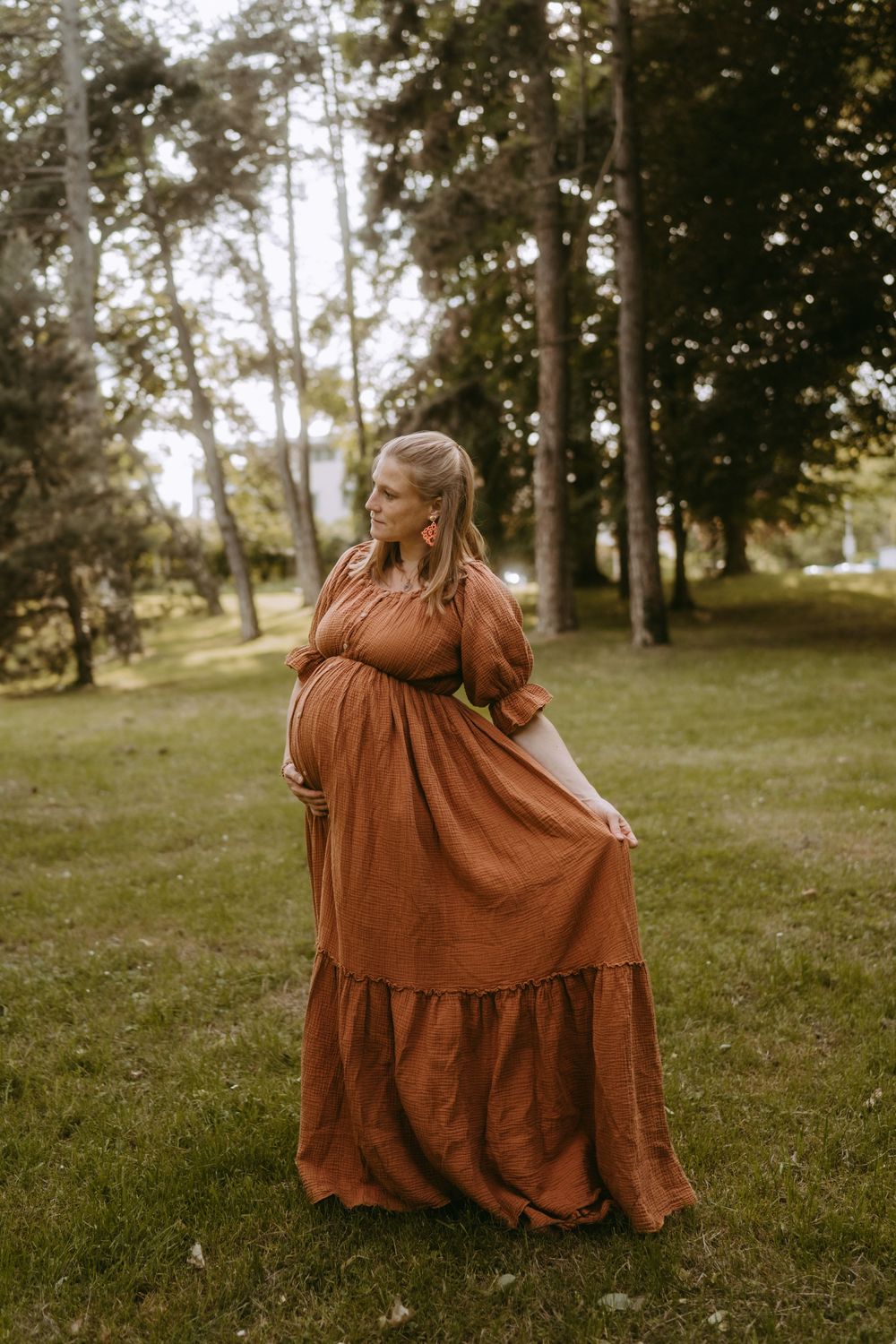 A pregnant woman in a rust orange maxi dress cradles her baby bump while standing in a green park.